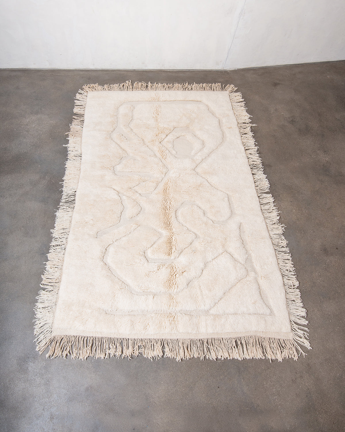 Berberlin x Malaika Raiss Beni M'rirt rug made of velvety cream sheep's wool with abstract woven design and natural wool tassels on all four sides, laying on concrete studio floor.